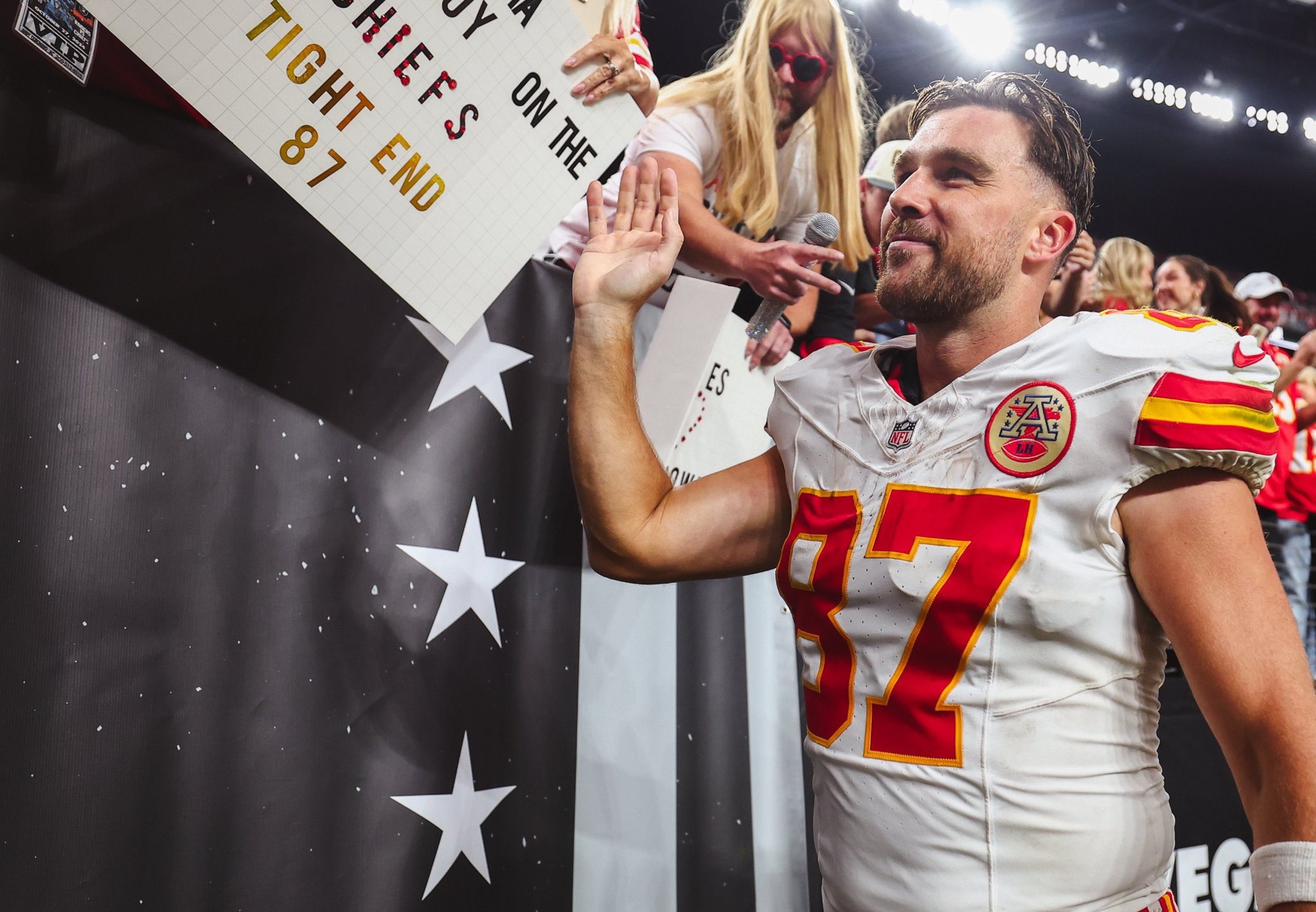 Travis Kelce smiling at a sign that says, “Karma is the guy on the